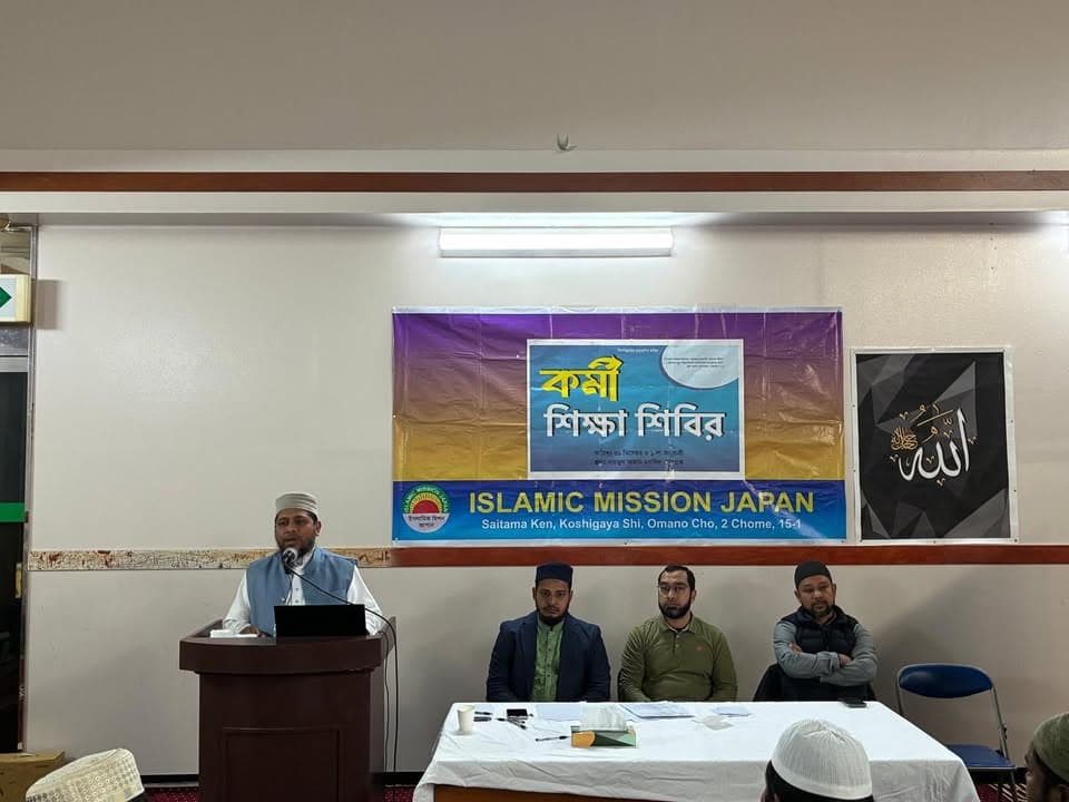 A man in a white cap speaks at a podium with a microphone in front of a wall banner reading "Islamic Mission Japan, Saitama Ken, Koshigaya Shi" and Bengali text, while three seated men in caps listen behind a table.
