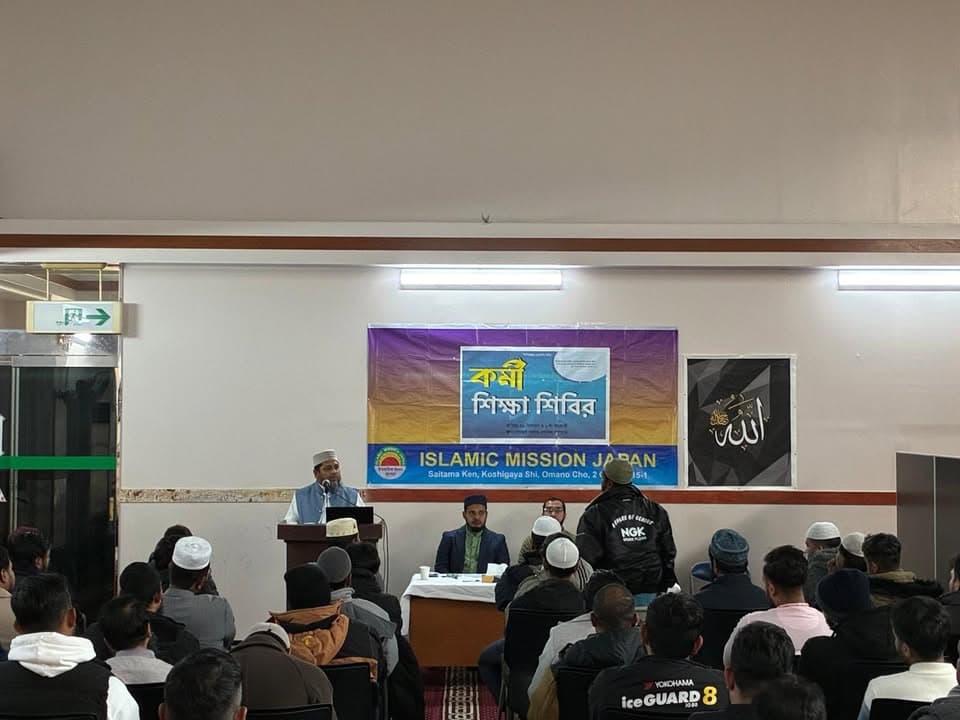 A speaker addresses a seated audience of men in a hall, standing at a podium beneath a Bengali-language banner reading "Workers Training Camp" and an "Islamic Mission Japan" sign, with an Allah calligraphy panel on the wall nearby.