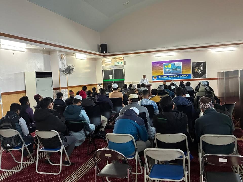 Rows of men sit on folding chairs inside a mosque hall, listening to a speaker at a podium beneath an "Islamic Mission Japan" banner written in Bengali and English.