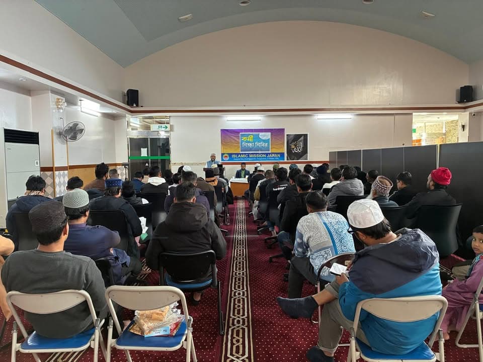 A large group of men wearing caps sit in rows of chairs inside a prayer hall, listening to two speakers seated at a table in front of an Islamic Mission Japan banner in Bengali and English.
