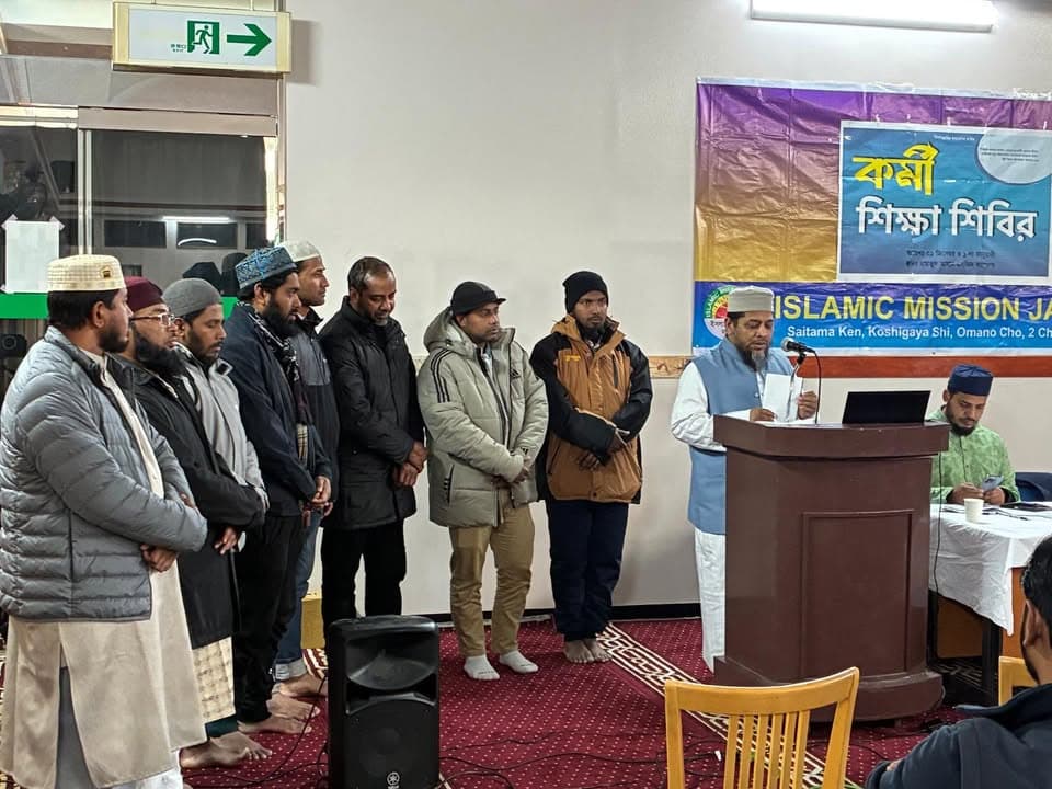A group of men stand in a line on a carpeted prayer hall while a speaker addresses them from a wooden podium, with a Bengali and English banner reading "Worker Training Camp, Islamic Mission Japan" hanging on the wall behind.