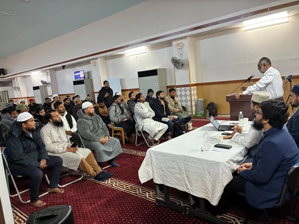 A man stands at a podium speaking into a microphone in a mosque hall, while rows of men sit on chairs listening, and two other men sit at a table with laptops in front of the podium.