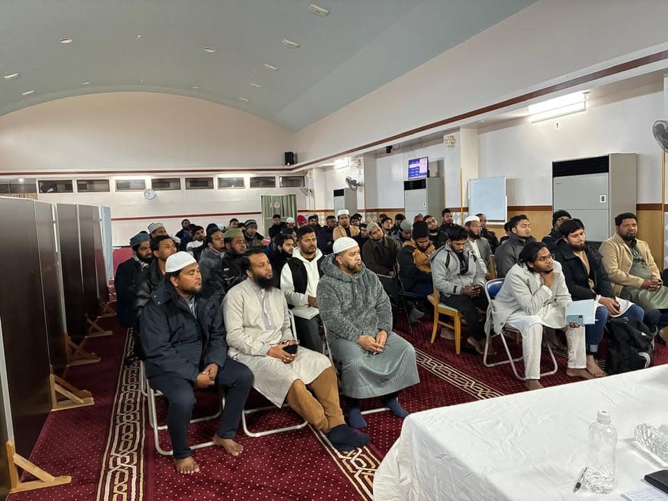 Rows of men, many seated in chairs and some on the floor, listen attentively inside a mosque hall with a red patterned carpet during a training session.