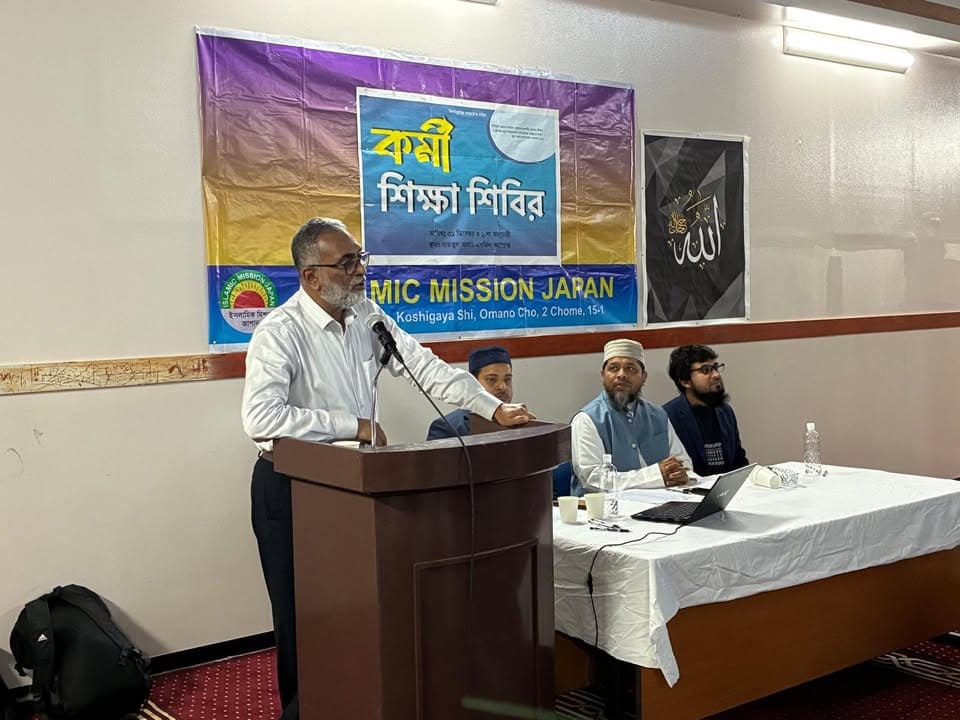 A man speaks at a podium microphone in front of a banner with Bengali text reading "Worker Training Camp" and "Islamic Mission Japan," while three men sit at a table listening.