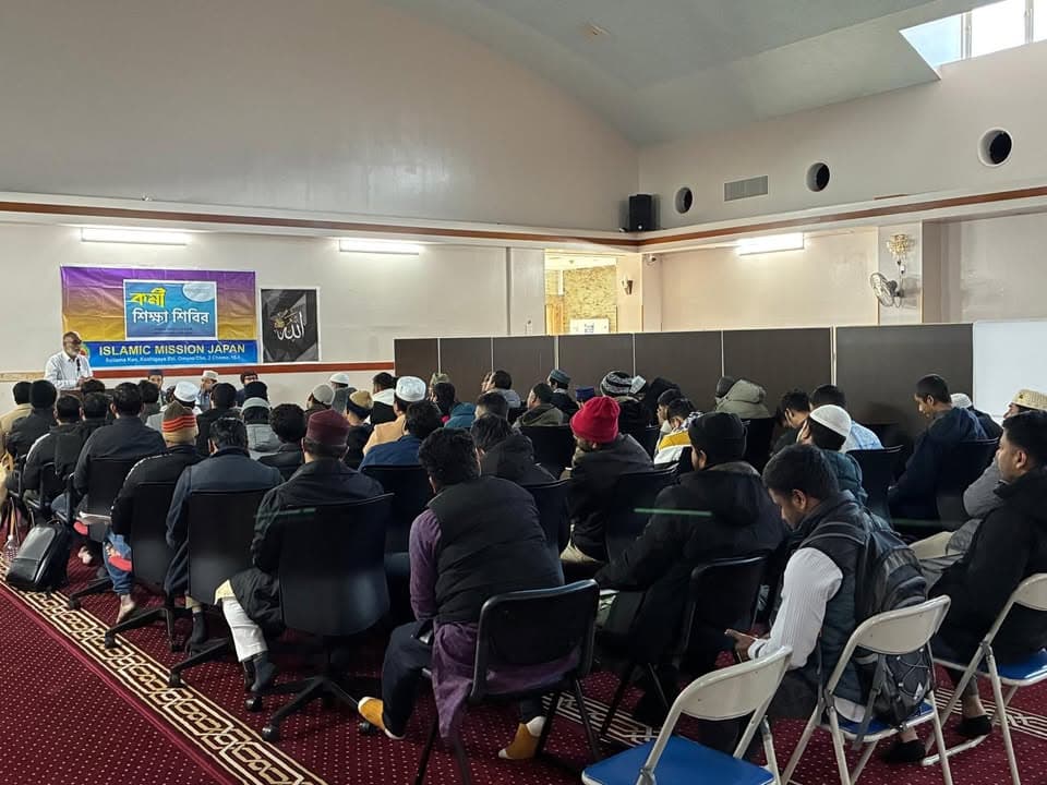 Rows of men in winter coats and caps sit on chairs facing a speaker at a lectern, with a banner reading "Islamic Mission Japan" and Bengali text hung on the wall behind him.