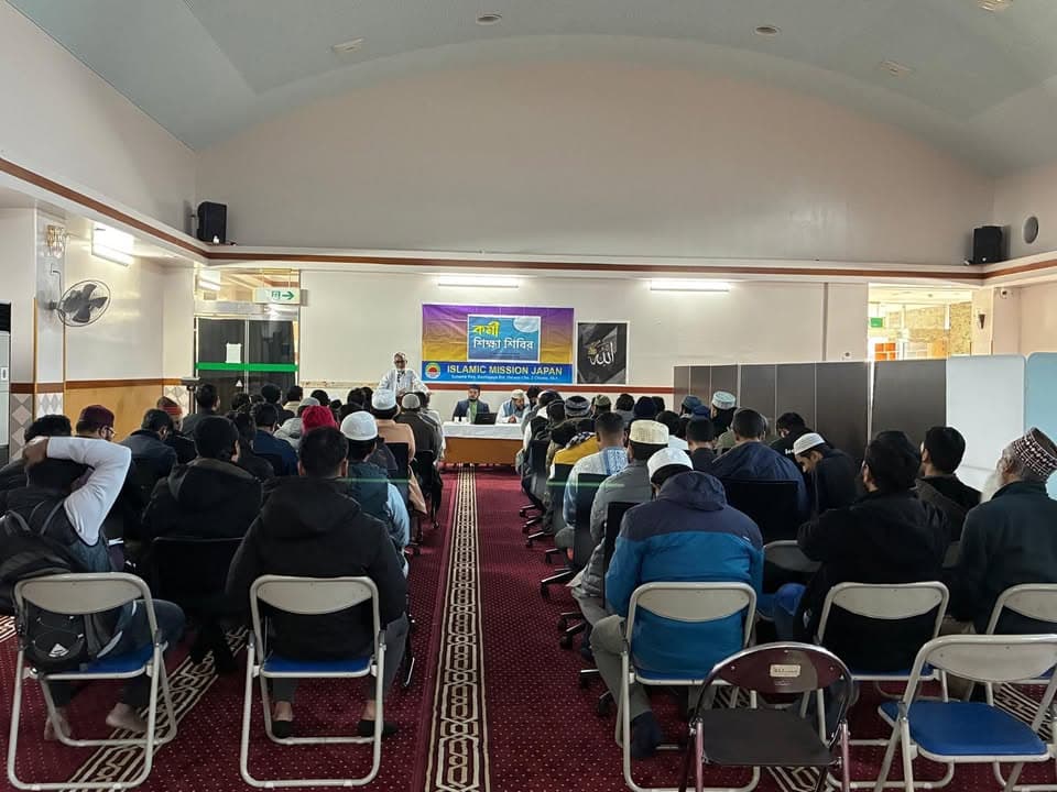Rows of men sit on chairs facing a front table with three speakers, beneath a banner reading "Islamic Mission Japan," inside a mosque hall with red patterned carpet.
