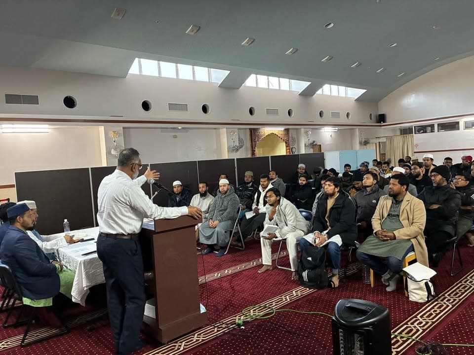 A speaker gestures from behind a podium microphone as he addresses a seated audience of men in a mosque hall with domed skylights and red patterned carpet, while others sit at a table nearby taking notes.