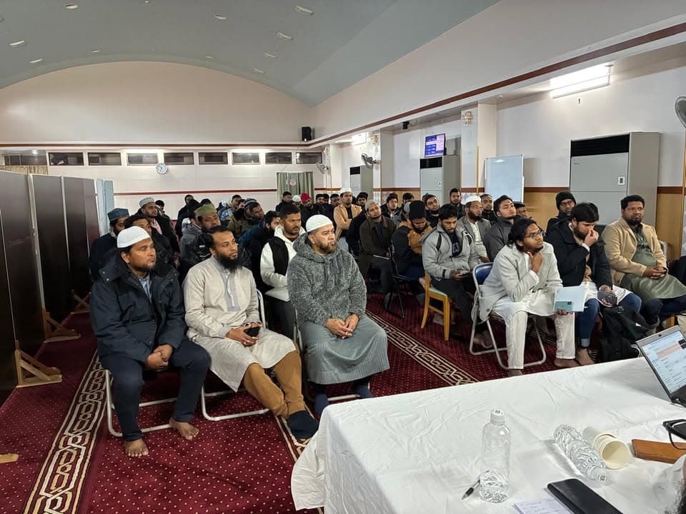 Rows of men in coats and prayer caps sit on chairs on a red patterned carpet inside a mosque hall, listening attentively to a speaker, with a table holding water bottles and a laptop in the foreground.