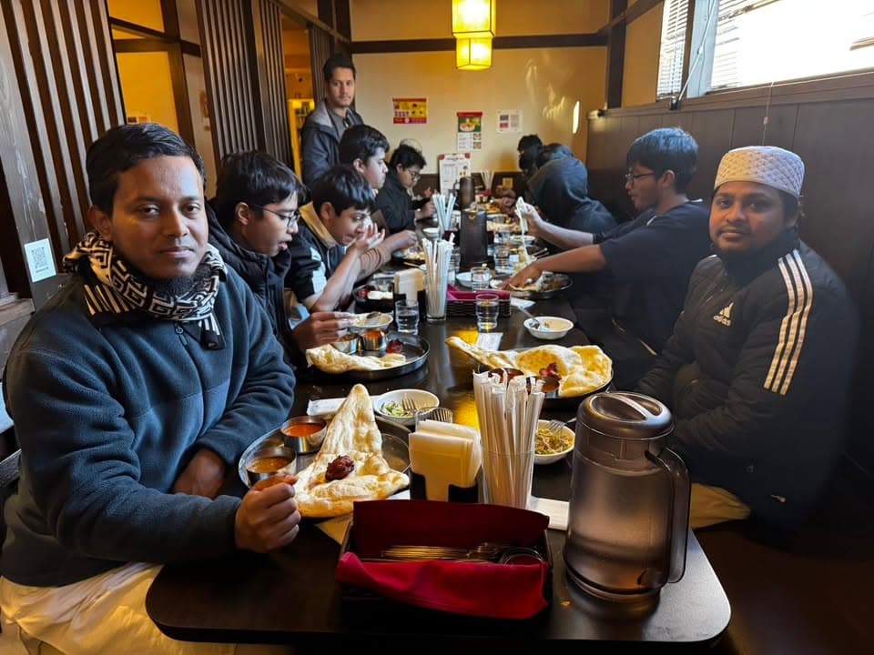 A group of men and boys sit along a long table at a restaurant, eating naan bread and curry dishes together, with two men looking toward the camera in the foreground.