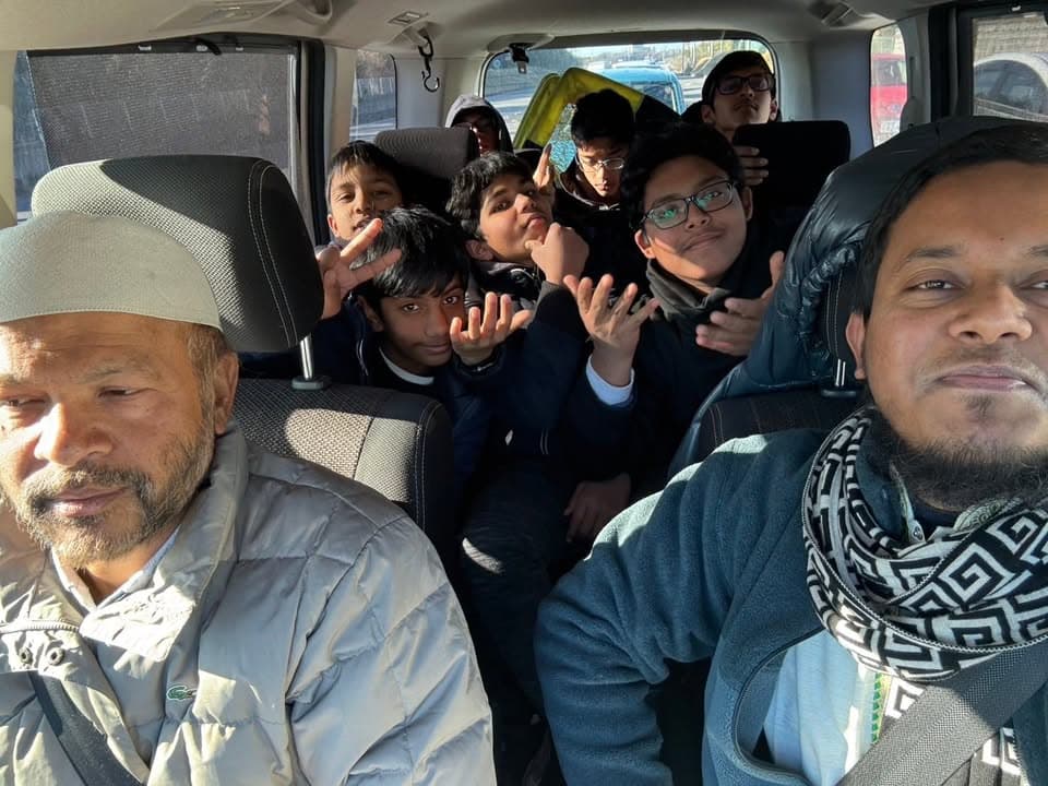 Two men and a group of boys ride together packed inside a van, with several boys making cheerful hand gestures toward the camera.