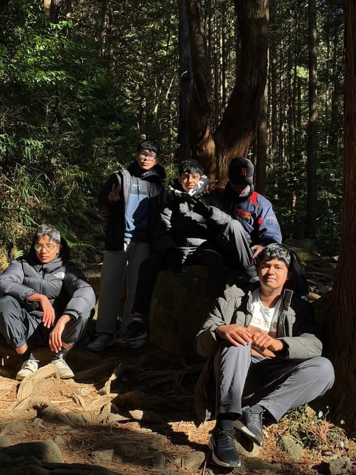 Five young men pose together on and around a large rock in a forest, surrounded by tall trees and sunlight filtering through the branches.
