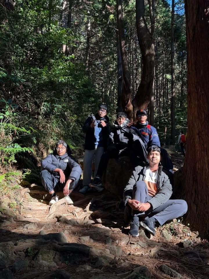 Five young men in warm outdoor clothing sit and crouch among tall trees and exposed roots on a wooded hillside, posing for a group photo in sunlight filtering through the forest.