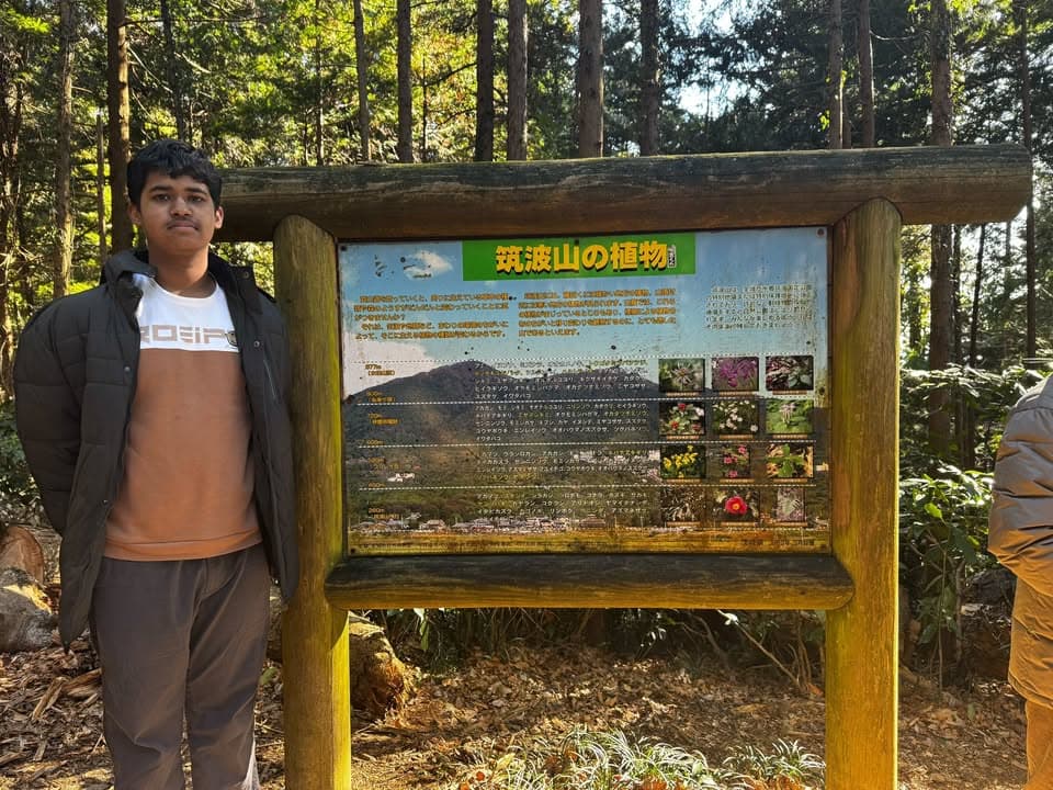A young man in a jacket stands beside a wooden information board titled "Plants of Mount Tsukuba," surrounded by tall trees in a forest.