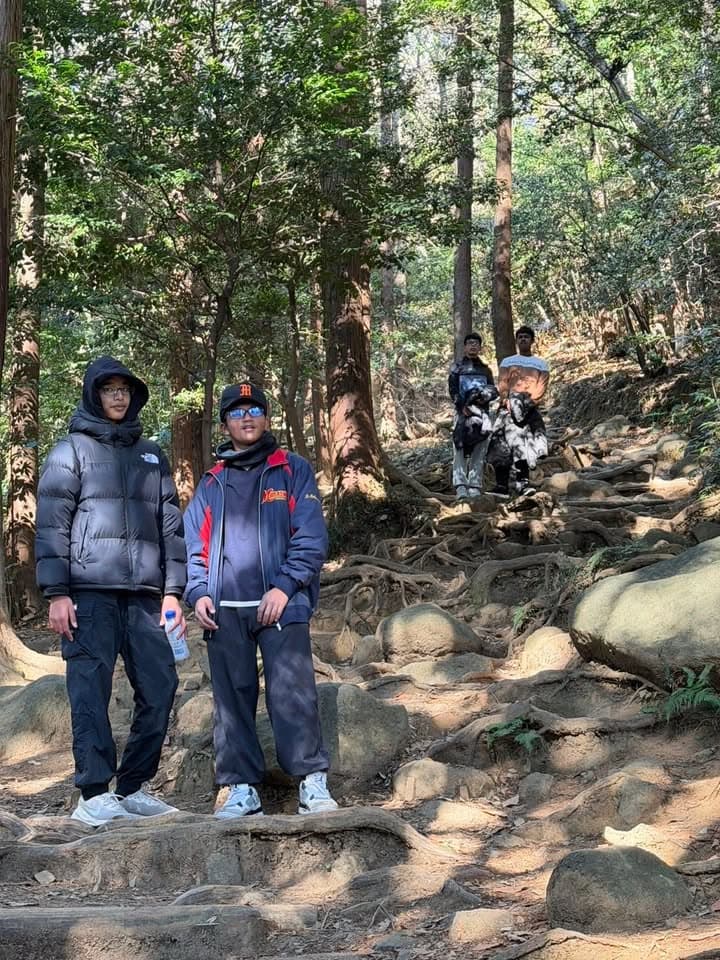 Group of young men in warm jackets pause on a rocky, root-covered forest trail while hiking up a wooded mountainside.