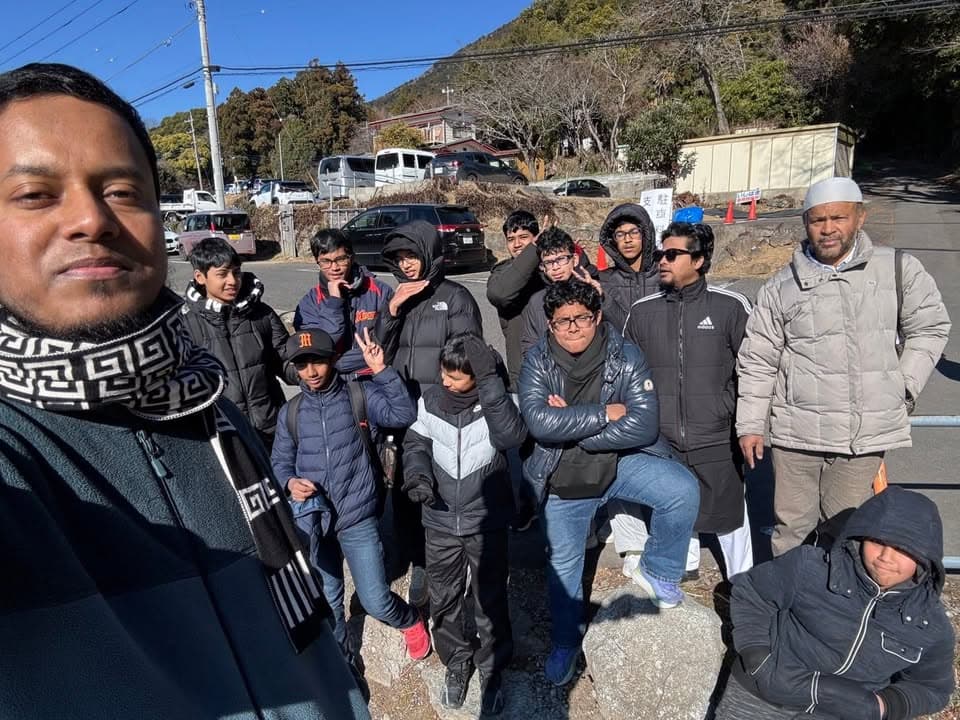 A group of men and boys in winter coats pose together for a selfie-style photo in an outdoor parking area, with cars, hills, and trees in the background.