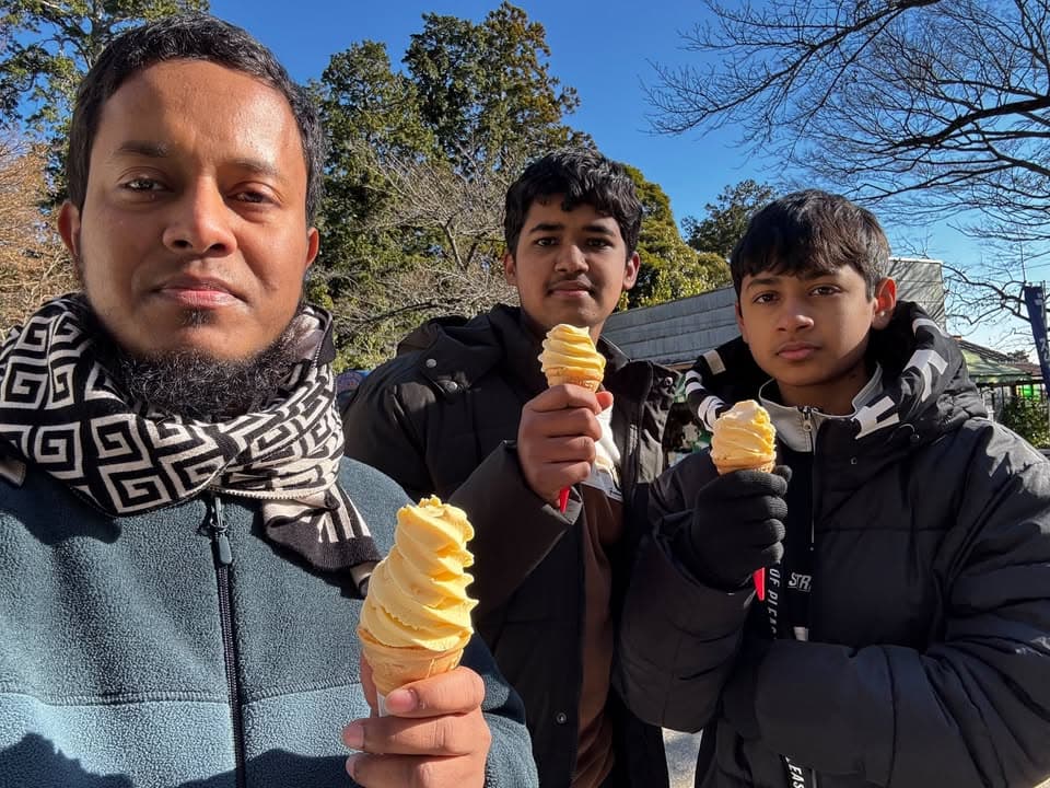 A man and two boys in winter jackets pose outdoors on a sunny day, each holding a soft-serve ice cream cone, with bare trees and a clear blue sky in the background.