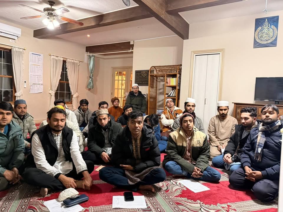 A group of men and youths, some wearing prayer caps, sit cross-legged on a red patterned carpet inside a room with wooden ceiling beams, listening attentively during an indoor gathering.