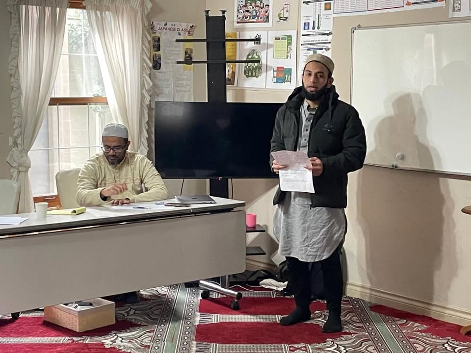 A young man wearing a cap and jacket stands reading from a paper as another man in traditional clothing sits at a desk taking notes nearby, in a room with a whiteboard, a large screen and a red patterned carpet.