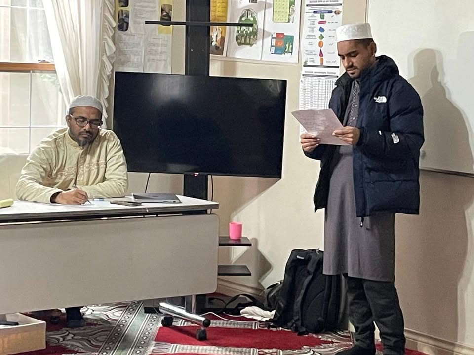 A man wearing a white cap and long white robe sits at a desk taking notes while another man in a black winter jacket stands and reads aloud from a paper, in a room with a wall-mounted TV screen and children's educational posters in the background.