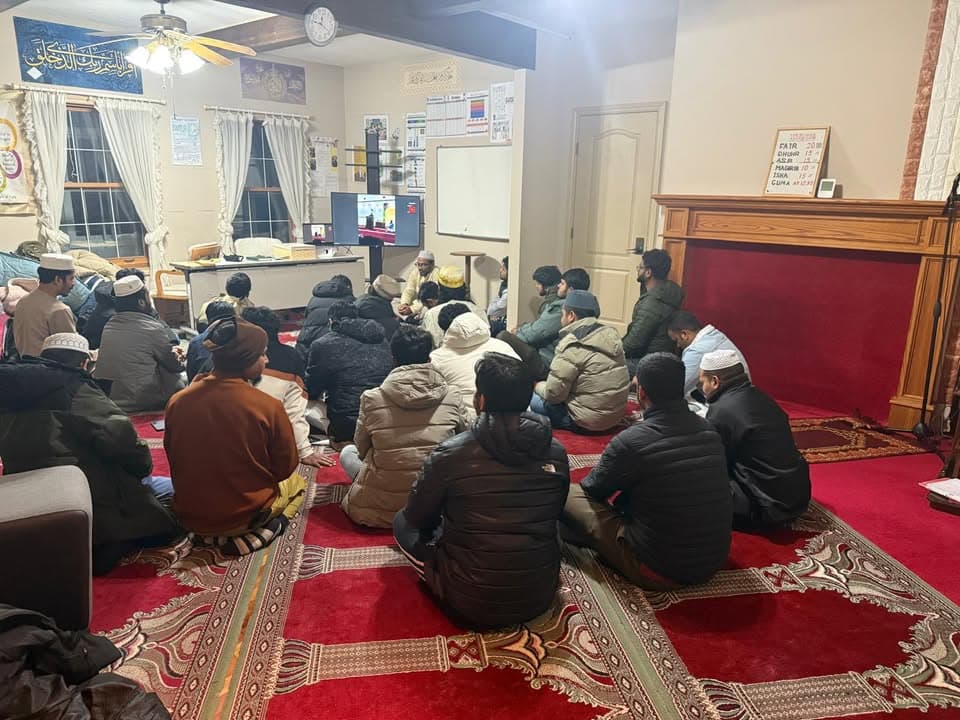 A group of men sit on a red carpeted prayer hall floor, listening to a speaker at the front near a whiteboard and television screen showing a video presentation.