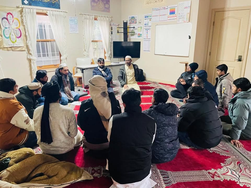 A group of men and young men sit in a circle on red carpets inside a mosque room, listening to a man in traditional white robes and a cap who is speaking to them.