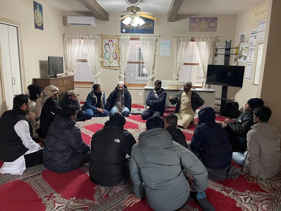 A group of men sit cross-legged in a circle on red patterned carpets inside a room with a masjid, listening to a man in a light robe and cap who is speaking near a table and TV screen.