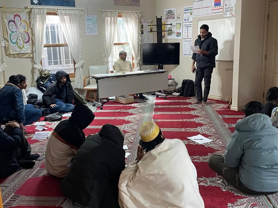 Men sit on a red patterned carpet in a mosque room, some wearing winter coats, listening as one man stands reading from a paper and another sits at a table at the front near a television screen.