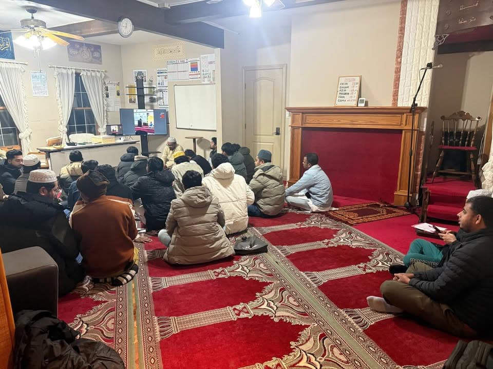 A group of men and boys in winter jackets sit cross-legged on red patterned prayer carpets inside a mosque hall, listening to a speaker near a whiteboard and television screen, with a wooden mantelpiece and prayer time board on the right.