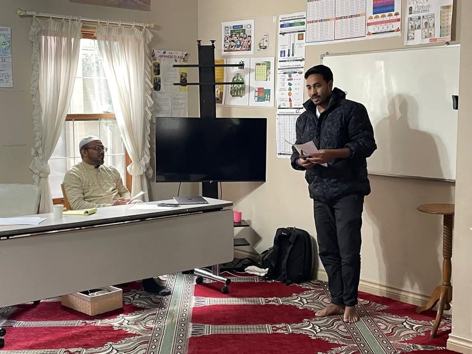 A man stands holding notes and speaking in a room with a whiteboard, TV screen, and educational posters on the wall, while another man in a white robe and cap sits and listens at a desk, on a red patterned carpet.