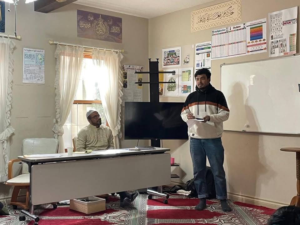 A man stands speaking and holding notes in a classroom-like masjid room, while another man in a kufi cap sits at a table listening, surrounded by educational posters, a whiteboard, and a red patterned carpet.