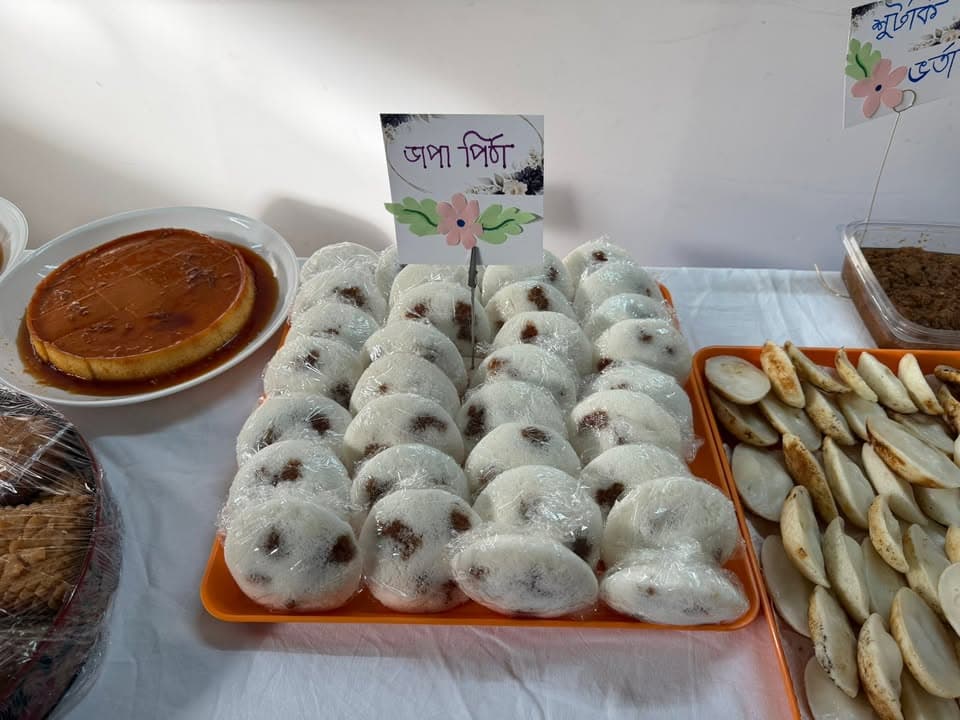 Trays of traditional Bangladeshi rice cakes (pitha) with a handwritten label, arranged on a table alongside caramel custard, coconut cookies, and other sweets.