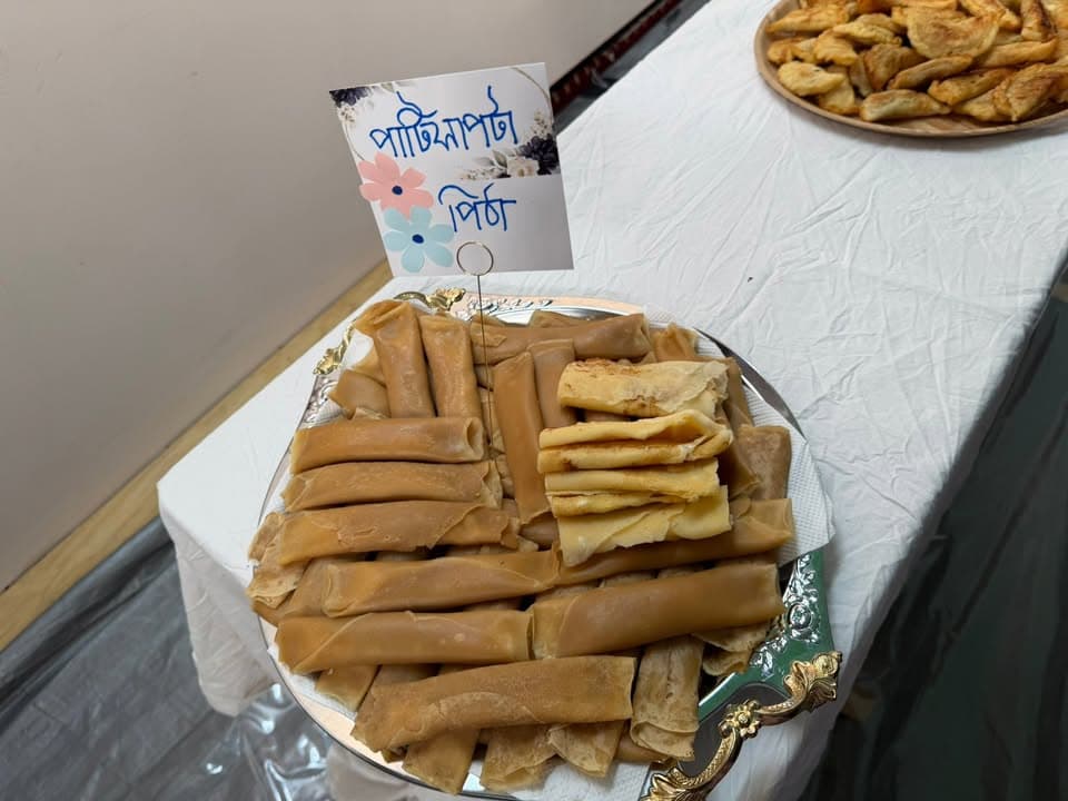 A silver tray on a white-clothed table holds rolled and folded Bengali pitha (rice cakes), with a small label card reading "Patishapta Pitha" in Bengali, and another plate of fried pastries visible in the background.