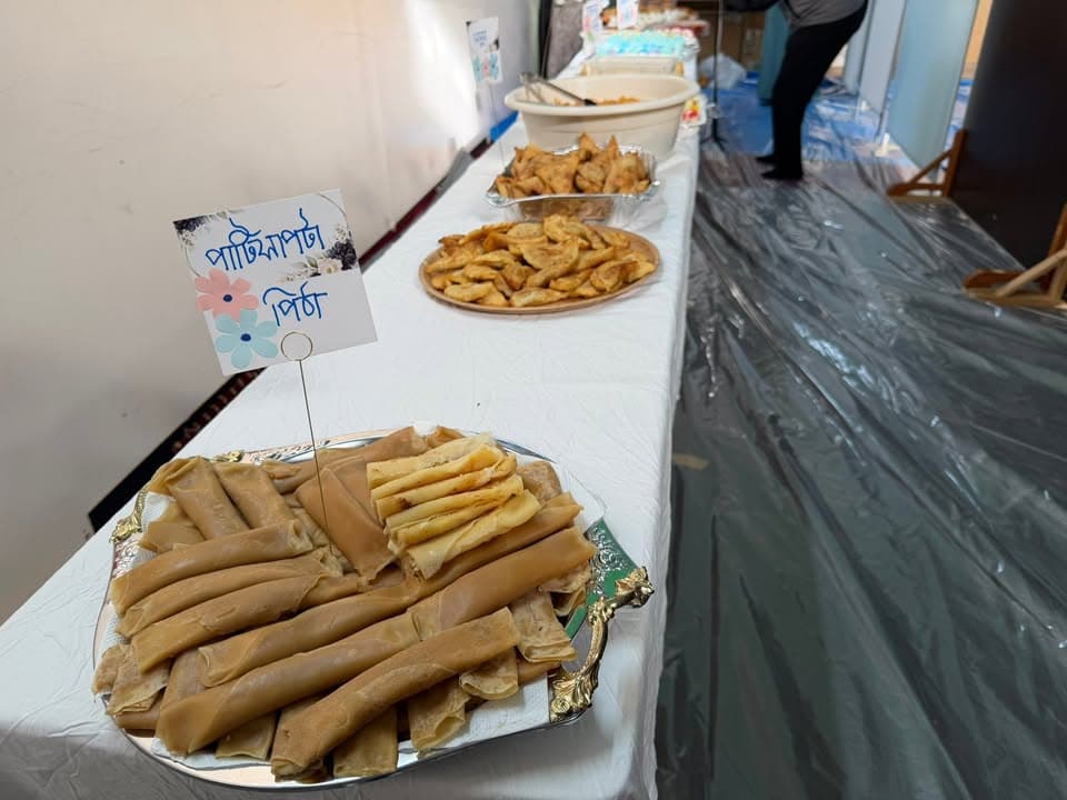 A long table set with trays of traditional Bangladeshi pitha rolls, fried snacks, and a Bengali-language name card, with a person standing near the far end of the table.