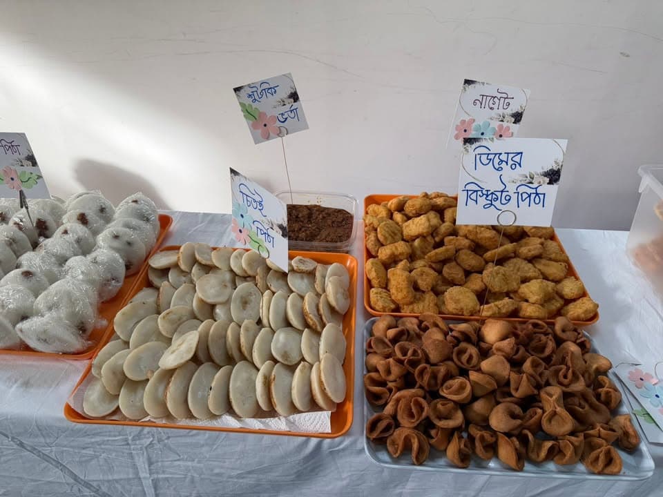 Trays of traditional Bangladeshi pitha and sweets, including rice cakes, fried dumplings, and gur (molasses), displayed on a table with handwritten Bengali name cards for each dish.