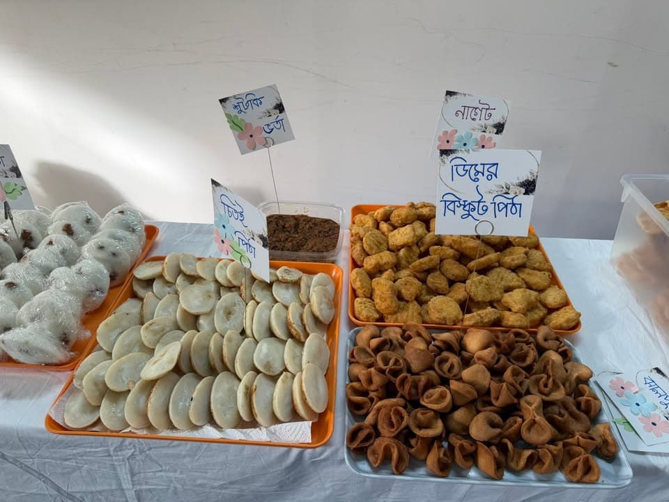 Trays of traditional Bangladeshi pitha (rice cakes), including chitoi pitha, gur-filled bhapa pitha, nakshi pitha, and fried pitha, displayed on a table with handwritten Bengali name cards.