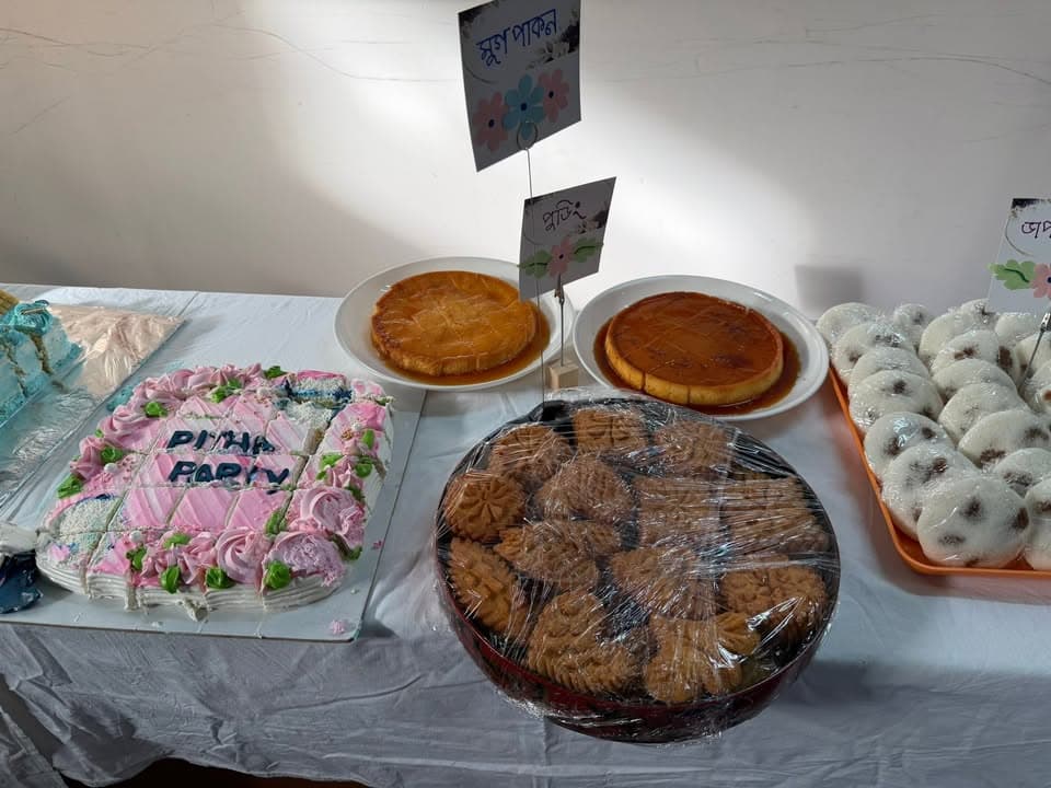 A table displaying various traditional Bangladeshi sweets and pitha, including a decorated cake reading "Pitha Party," round caramel-glazed puddings, plastic-wrapped cookies, and coconut-filled white rice cakes, each with small Bengali-language name cards.
