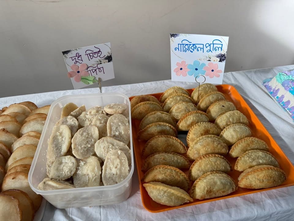 A table displaying trays of traditional Bengali pitha (rice cakes), including flat pancake-style pithas, coconut-filled half-moon pastries, and a container of dudh chitoi pitha, each labeled with handwritten Bengali name cards.