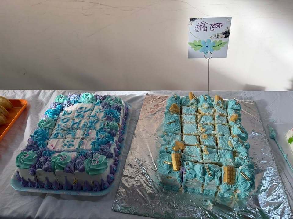 Two decorated sheet cakes with blue and purple frosting, cut into squares and topped with wafer sticks, displayed on a table with a small Bengali-language sign reading "পেস্ট্রি কেক" (pastry cake) placed between them.