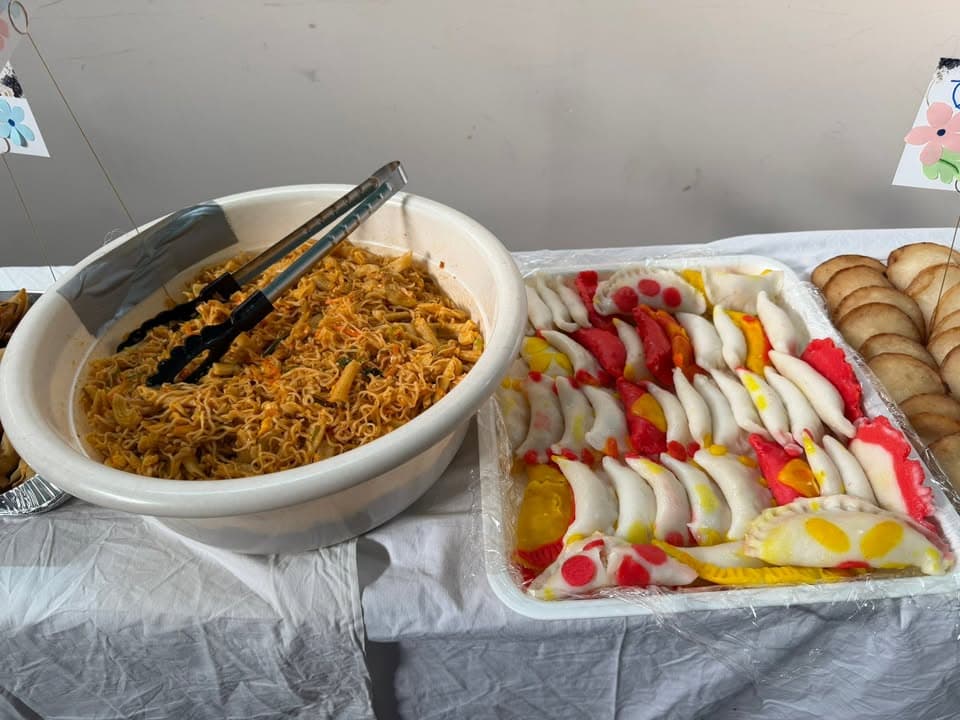 A large bowl of fried noodle salad with tongs sits beside a tray of colorful stuffed dumpling-style pitha and fried breads, all displayed on a cloth-covered table.