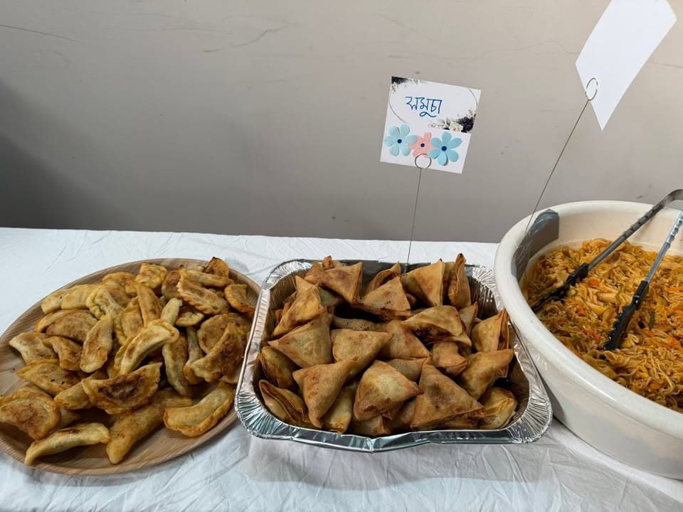 A buffet table displaying fried pastries, samosas in a foil tray with a Bengali-labeled sign, and a bowl of noodle-style pilau with serving tongs.