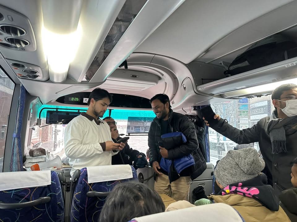 Passengers stand and sit inside a tour bus, with men chatting near the front while snow-covered streets are visible through the windshield.