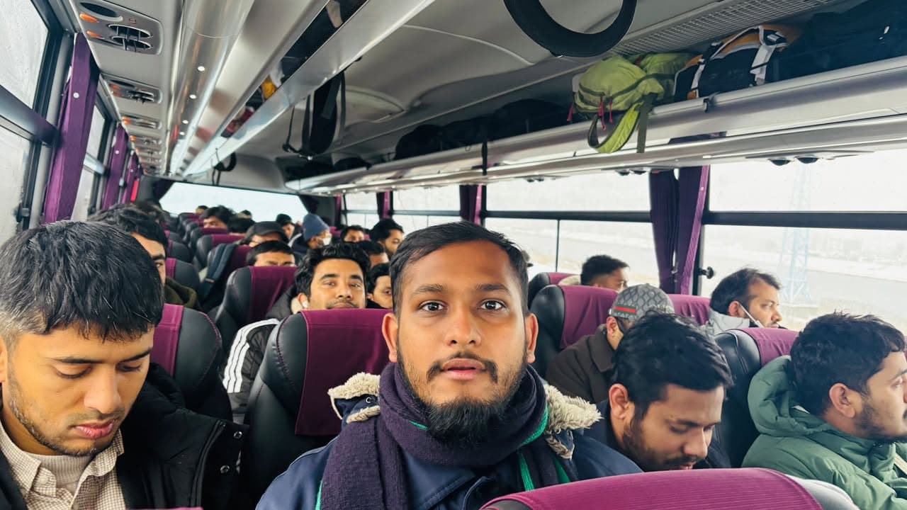 Passengers wearing winter coats and scarves sit closely together in a full charter bus, with backpacks stowed in the overhead racks, as one man in the foreground looks toward the camera.