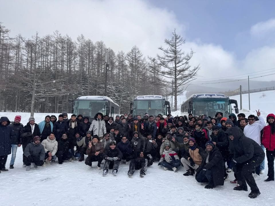 A large group of bundled-up participants pose together in the snow in front of three tour buses, surrounded by snow-covered trees.