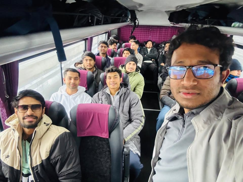 A group of men in winter jackets sit together on a charter bus, several looking toward the camera as one man in the foreground takes a selfie.