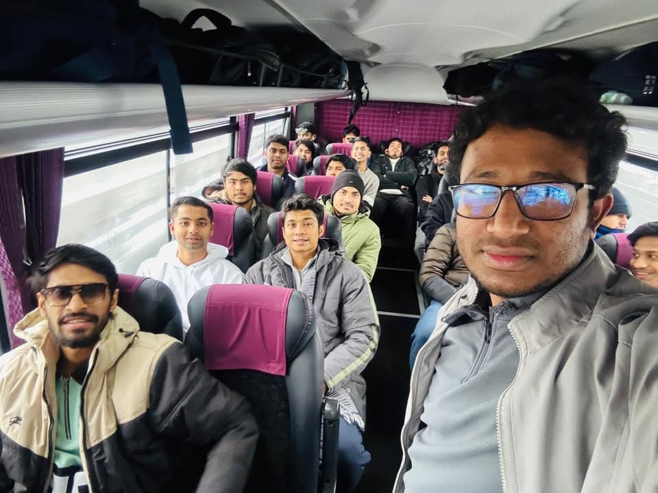 A group of young men in winter jackets sit in the seats of a charter bus, with one man in the foreground taking a selfie photo including many passengers behind him.