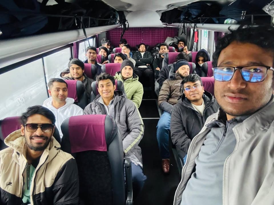 A group of men in winter coats sit in the seats of a charter bus, smiling and looking toward the camera as one man in the foreground takes a selfie.