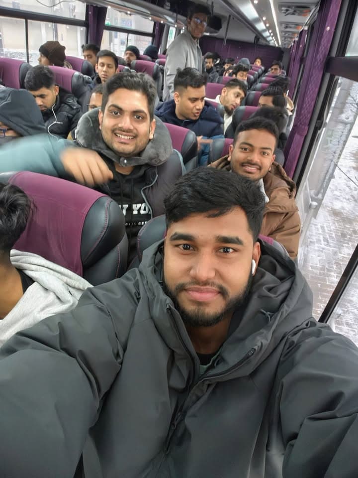 A group of young men in winter coats sit together on a charter bus, smiling for a selfie photo while other passengers fill the seats behind them, with snow visible outside the windows.