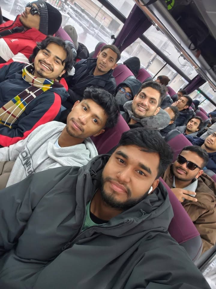 A group of men in winter coats sit together on a bus, smiling and posing for a group photo while traveling.