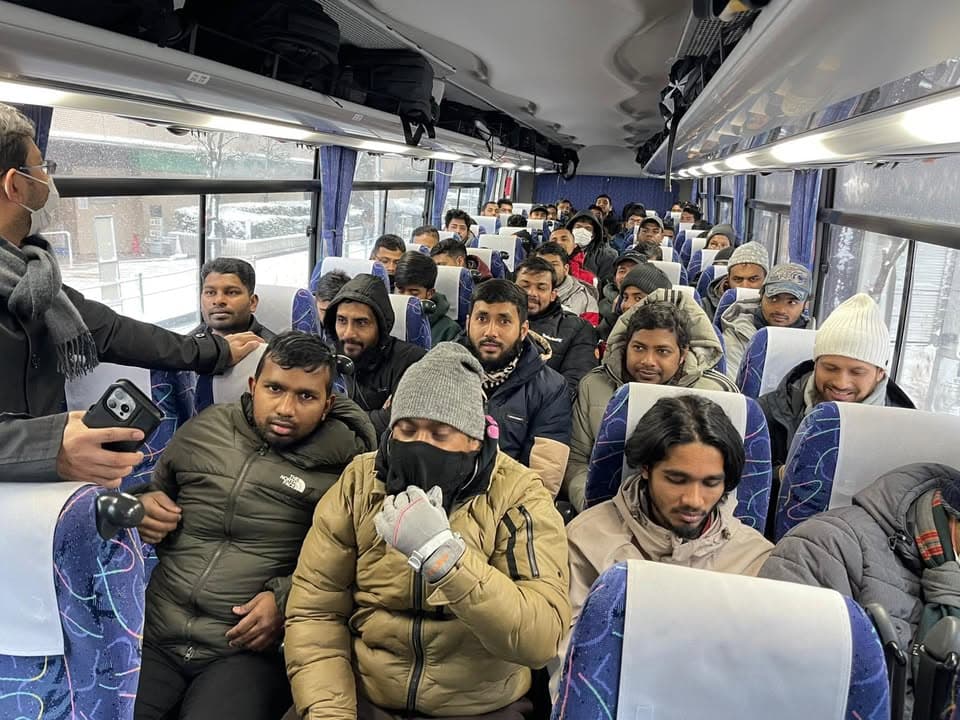 Passengers in winter coats and hats sit in the seats of a chartered bus, with snow visible through the windows outside, while a man wearing a mask and scarf stands in the aisle addressing them.