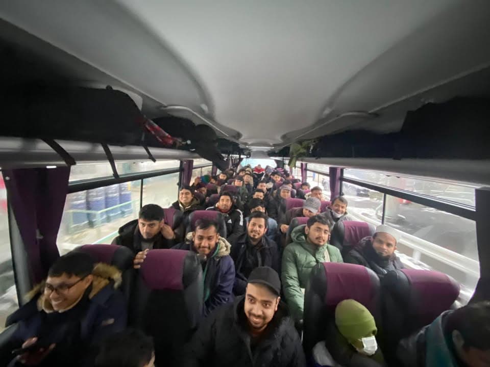 A group of men in winter coats sit together in rows of purple seats on a charter bus, smiling and turning toward the camera.